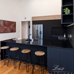 A photo of a kitchen island with 4 chairs a fridge and a painting on the wall.