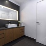 A photo of a counter top with a sink and a plant on it. There is a door on the left and a mirror above the sink.