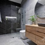 A photo of a bathroom area, with a shower in the background, toilet and large sink, with large round mirror over it.