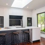 A photo of a kitchen area, with a centre island table and lush greenery in the background, outside the window.