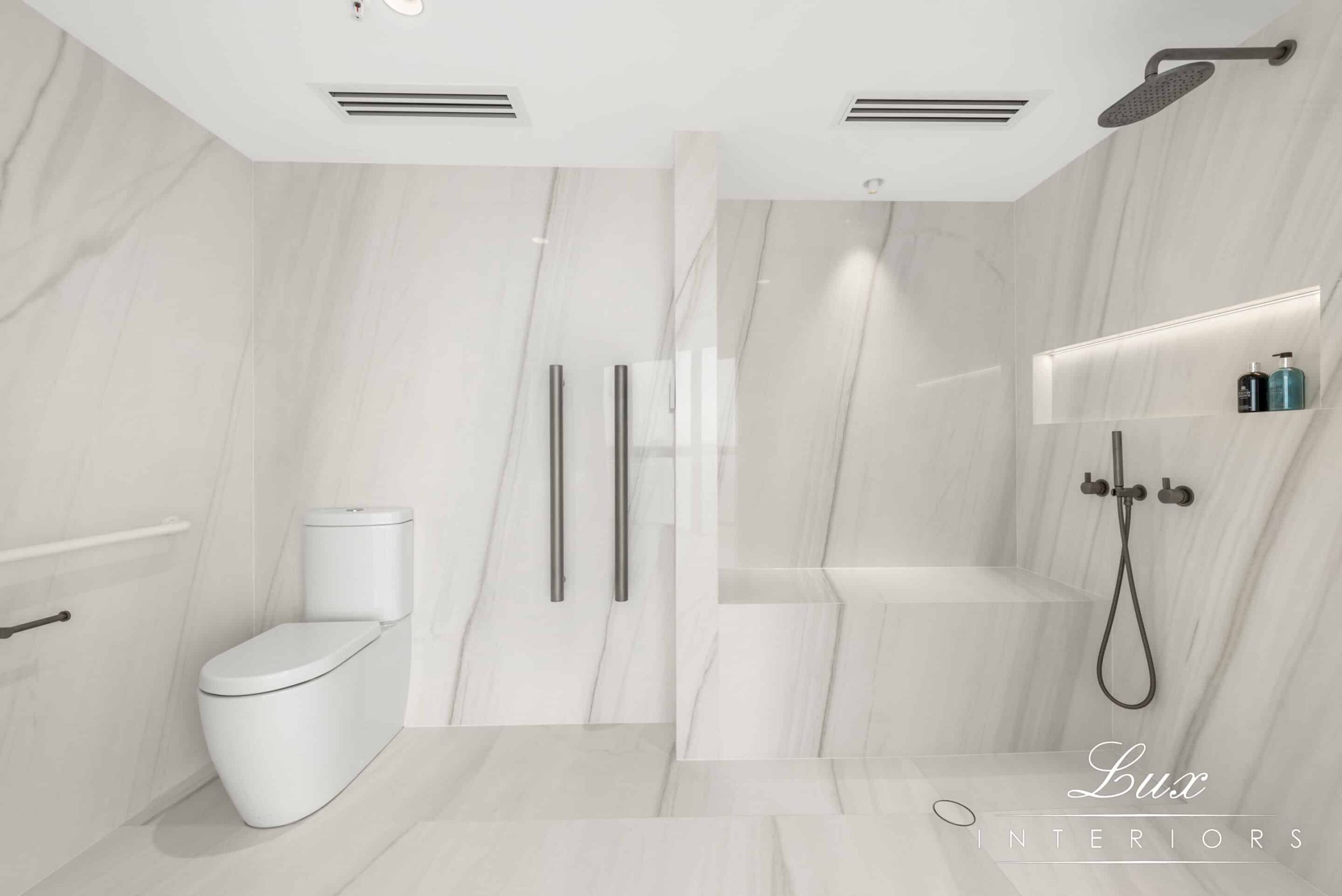 A white-marble bathroom, with toilet on the side and shower on the right side. There is a seated area for people needing wheelchair access to sit down whilst showering.