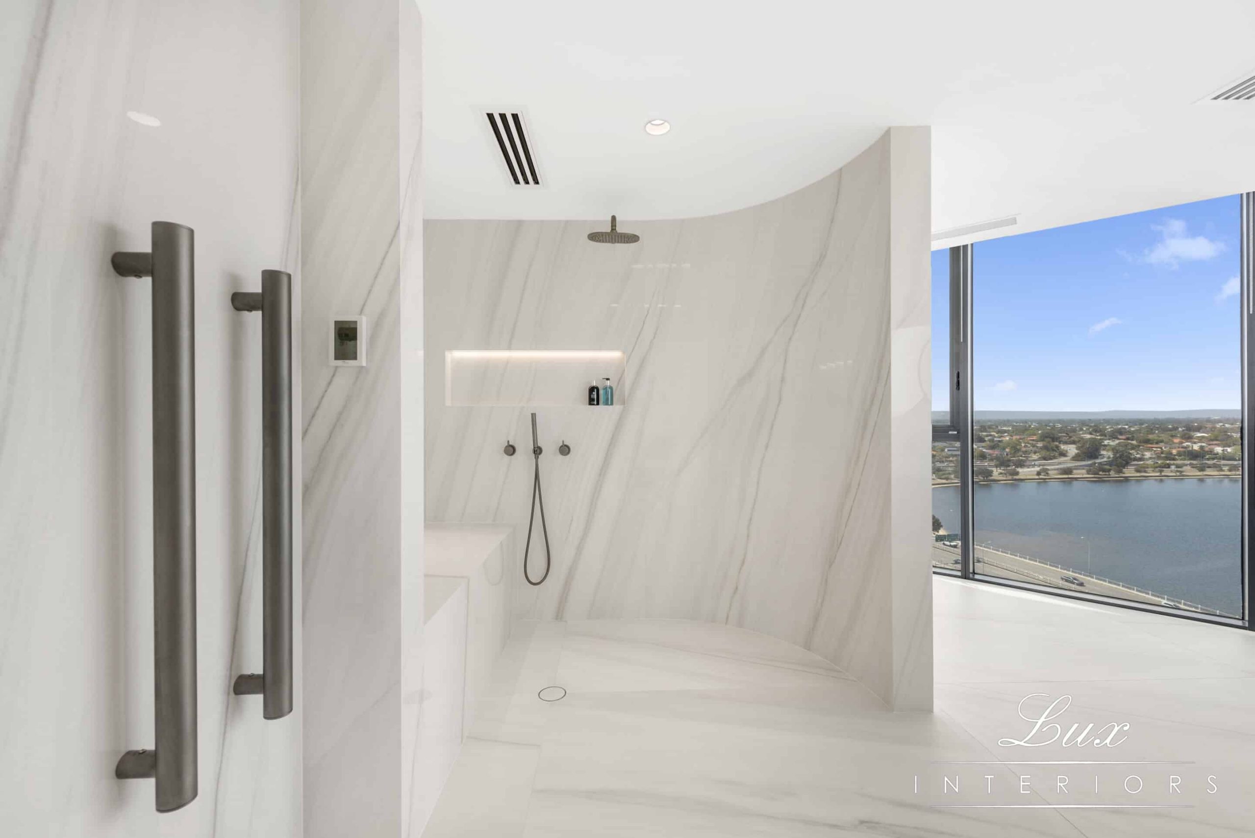 A white-marble bathroom, with toilet on the side and shower on the right side. There is a seated area for people needing wheelchair access to sit down whilst showering. There are views of the swan river in the background.