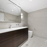 A photo of a large bathroom area, with a rectangular tap and large mirror, toilet in the background.