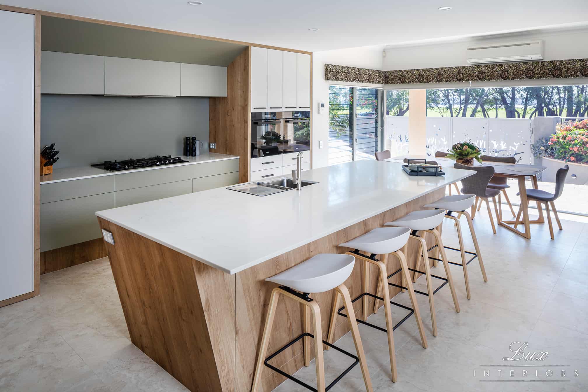 A photo of a freshly renovated kitchen area, with a white kitchen island, high chairs and large glass window to see outside.