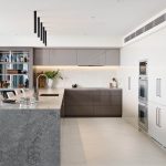 A photo of a kitchen area, with stone bench tops, white cabinets and a glass liquor cabinet in the back left.