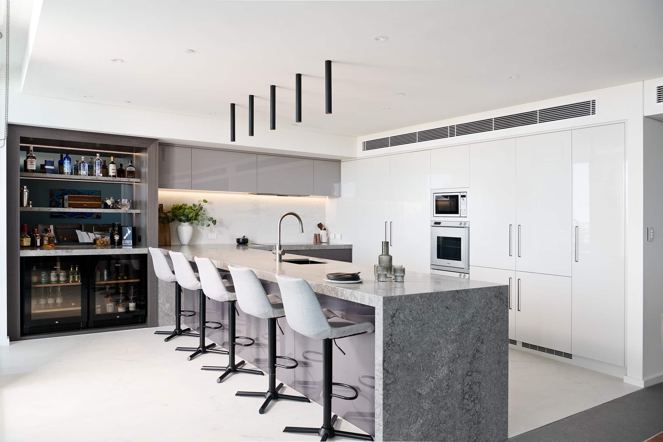 A photo of a kitchen area with stone benchtops and white stools.