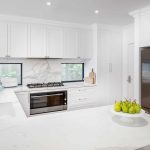 A photo of a kitchen, with white bench tops, a dark fridge and pears on a white plate.