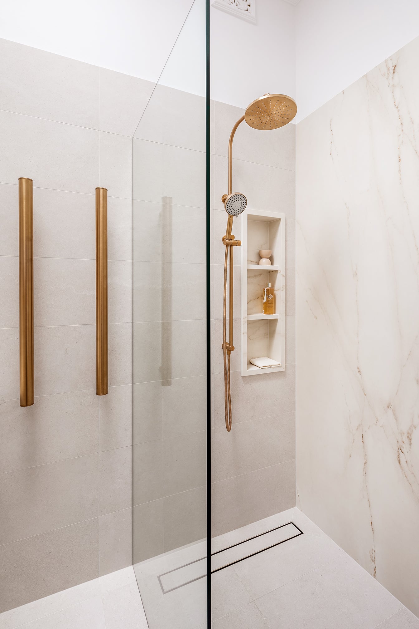 A photo of a shower, with a gold shower head and cream marble walls.