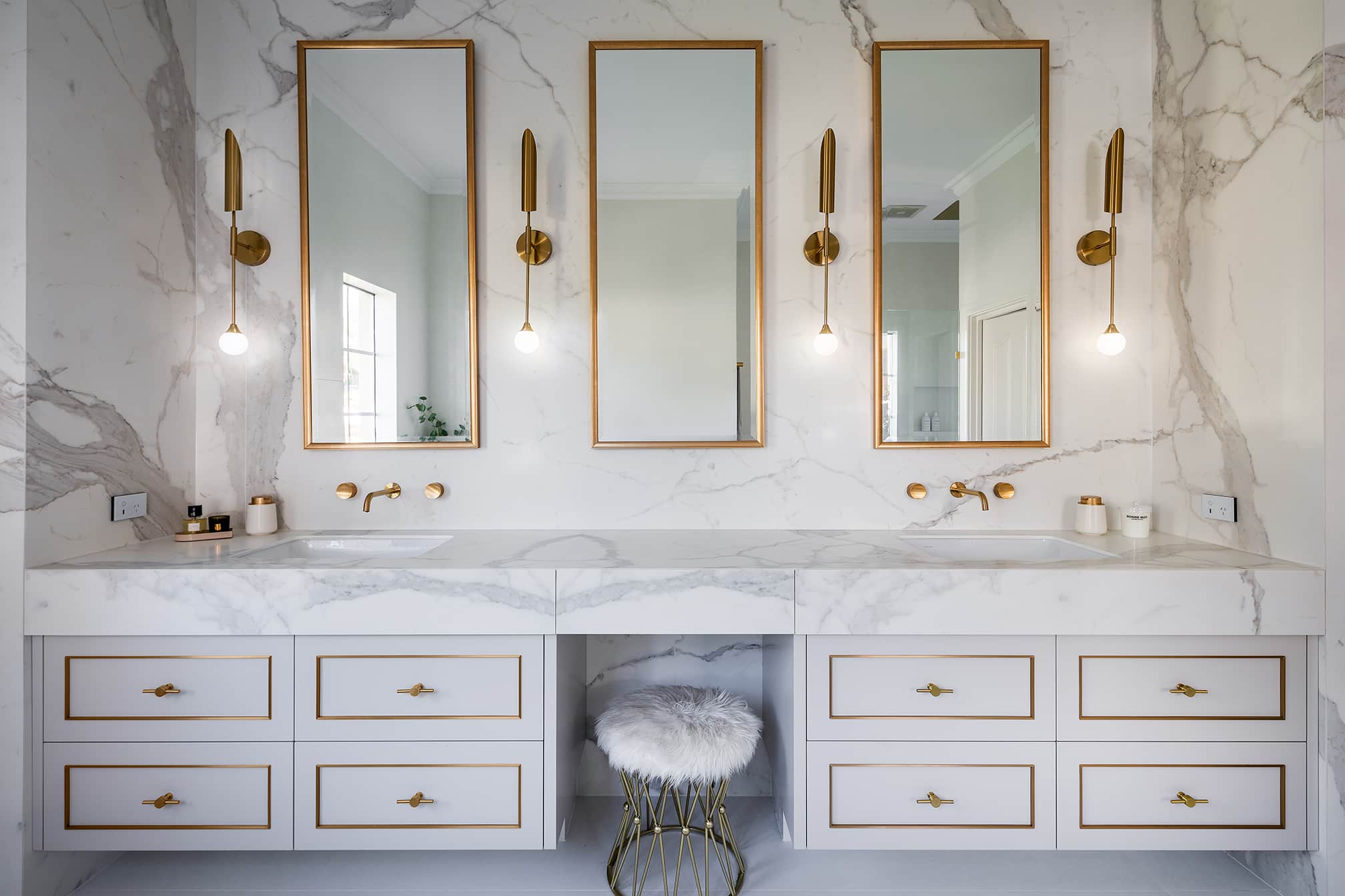 A photo of a bathroom, with white marble walls and 3 large, rectangular mirrors in the middle. There are two, dual sinks.