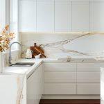 A photo of a white luxury kitchen with marble backsplash, a sink and stove in the background.