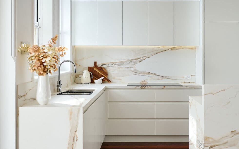 A photo of a white luxury kitchen with marble backsplash, a sink and stove in the background.