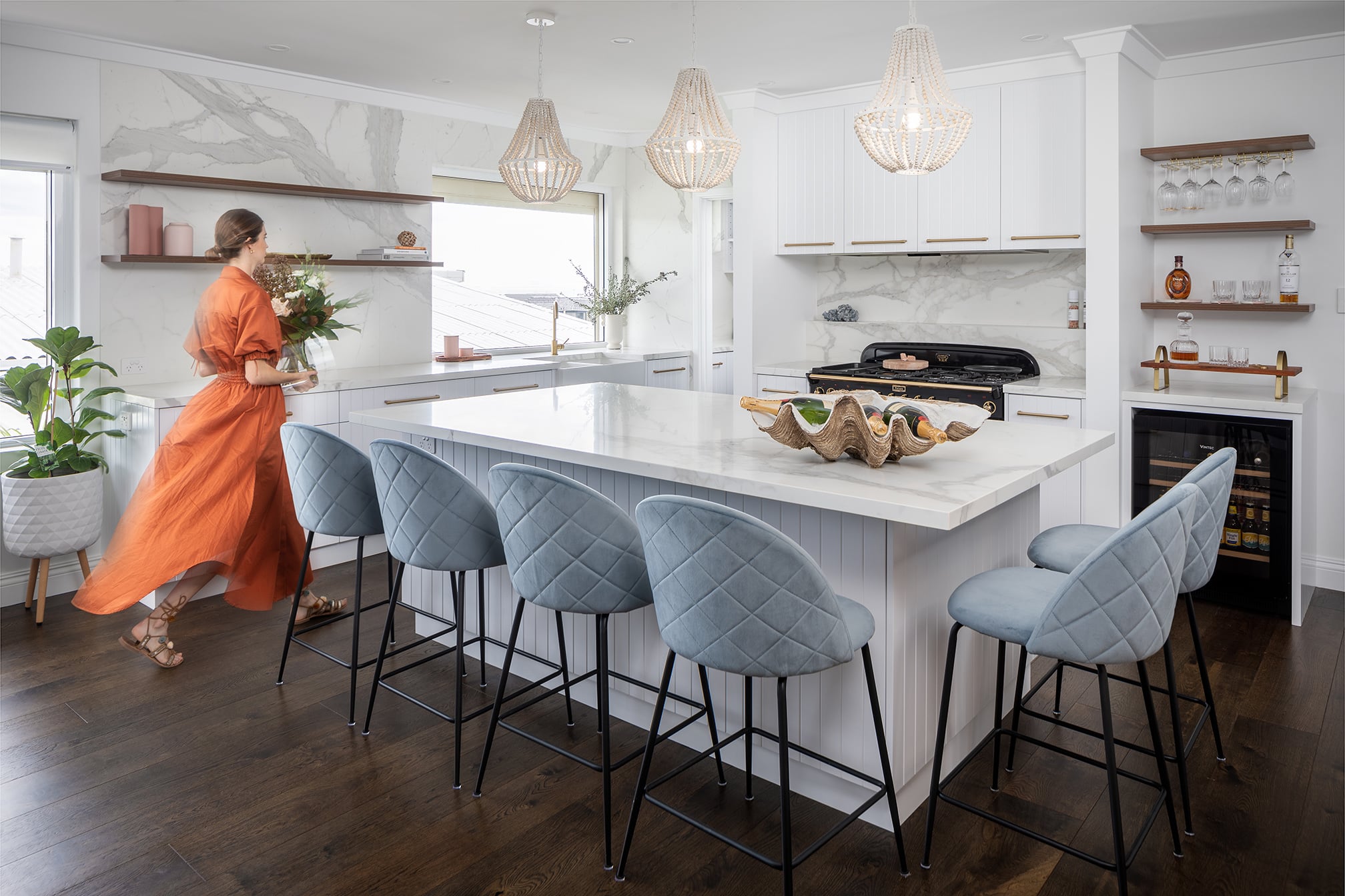 A photo of a centre island table, in a kitchen, with a woman walking to the side, wearing an orange dress.