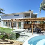 A photo of the back yard of a house, with lush green lawns, a wood patio and a rounded pool, filled with pristine blue water.